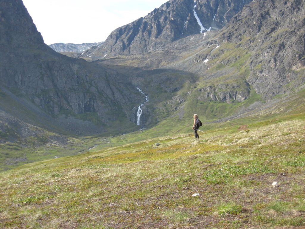 A lone hiker ascends a grassy mountain slope with towering peaks and a waterfall in the distance in Mat-Su, Alaska