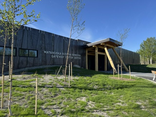 A modern visitor center with a wooden facade and large windows, surrounded by green grass and young trees in Mat-Su, Alaska