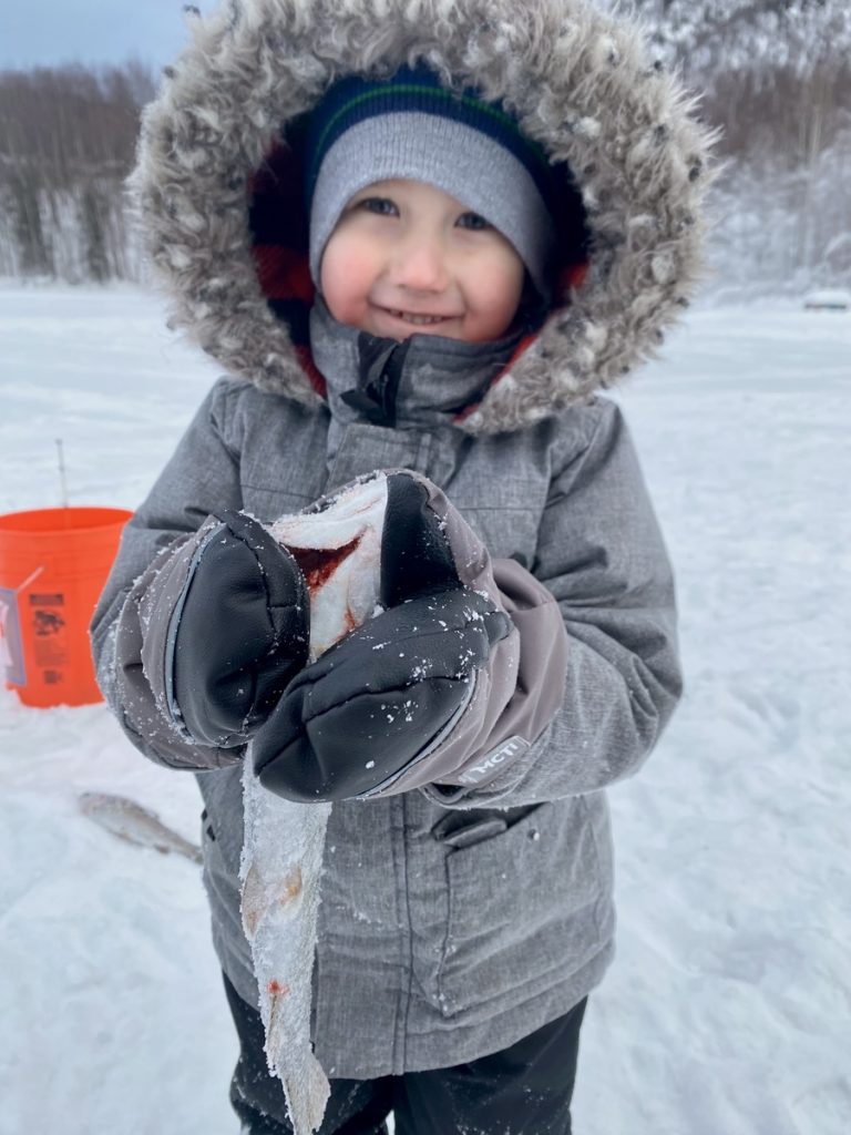 A young child, bundled up in a warm winter coat with a fur-lined hood, smiles while holding a piece of ice in their gloved hands in Mat-Su, Alaska