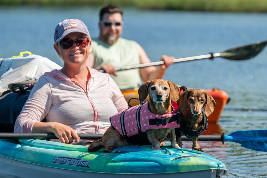 A woman in a kayak with two dachshunds wearing life jackets paddles on a calm lake with another kayaker in the background in Mat-Su, Alaska.