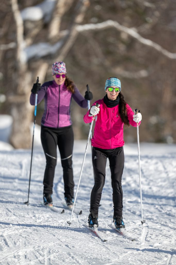 Two women cross-country skiing on a snowy trail, dressed in colorful winter gear and smiling in Mat-Su, Alaska