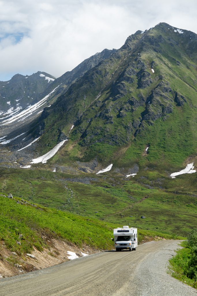 A white RV travels on a winding dirt road through a lush green valley with snow-capped mountains in the background in Mat-Su, Alaska