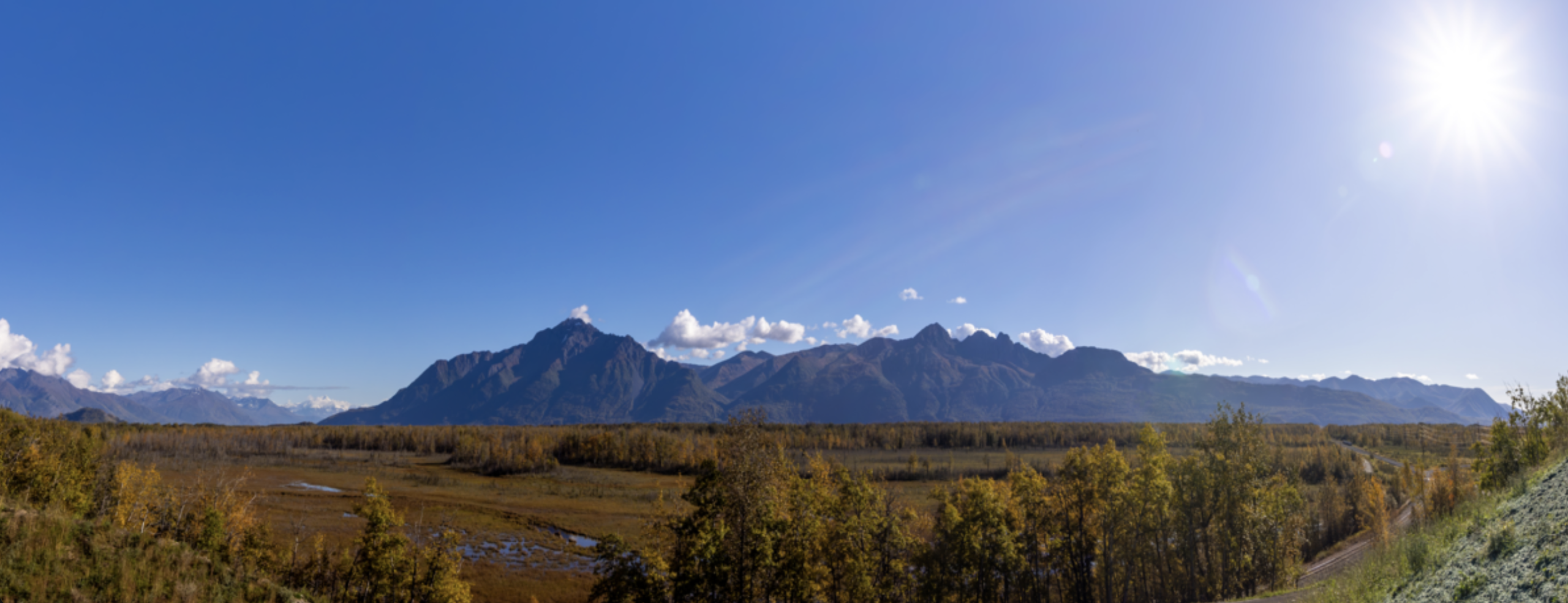 A panoramic view of a vast landscape with a river meandering through a valley, surrounded by colorful autumn trees and distant snow-capped mountains under a clear blue sky in Mat-Su, Alaska