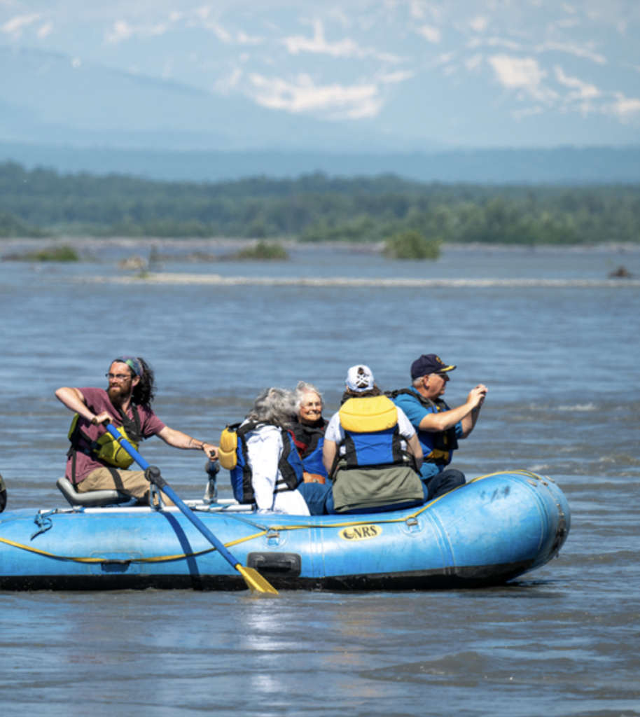 A group of adventurers in a blue raft navigate a river surrounded by lush greenery and distant mountains in Mat-Su, Alaska