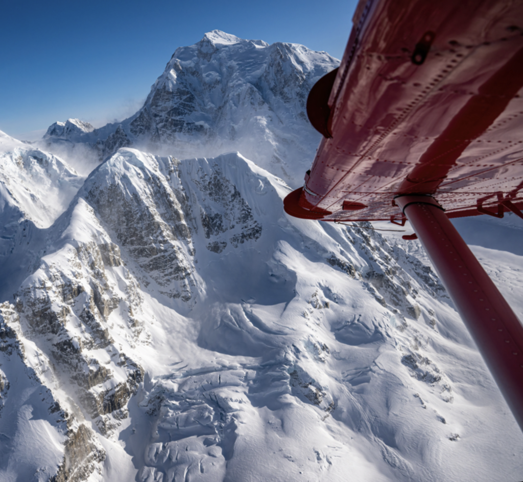 A red helicopter hovers near the peak of a snow-covered mountain range in Mat-Su, Alaska