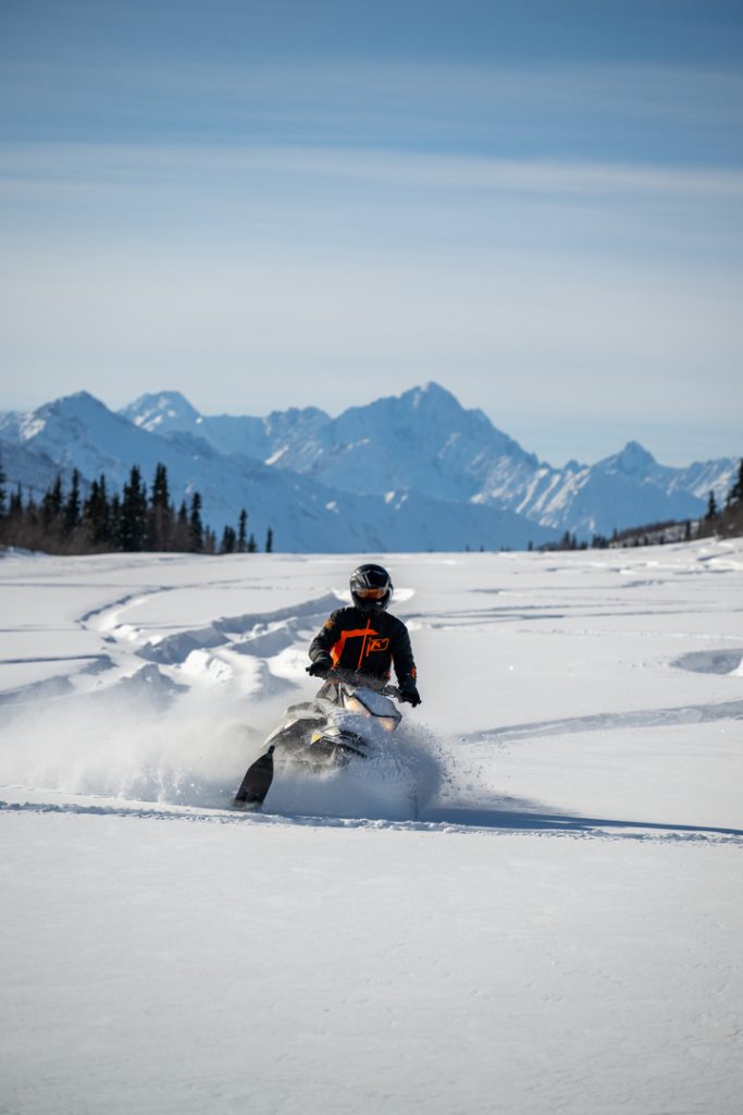 A snowmobiler in an orange jacket and black helmet speeds across a vast, snow-covered plain with majestic snow-capped mountains in the distance in Mat-Su, Alaska