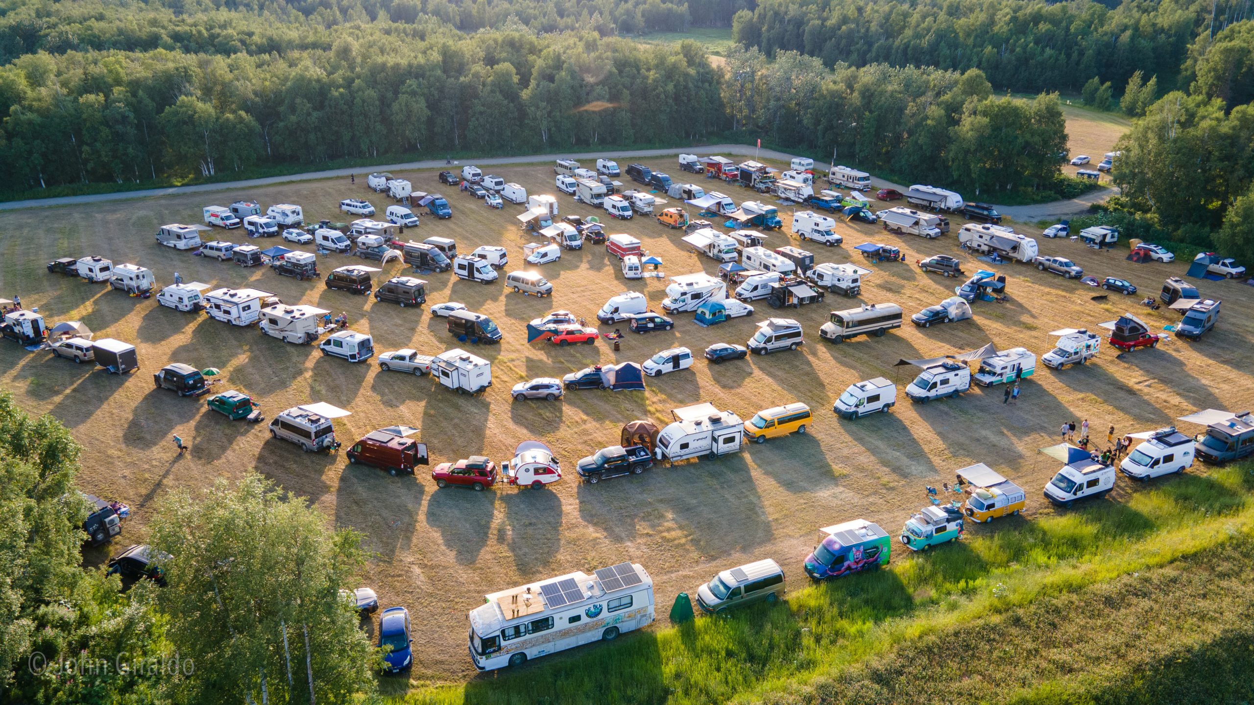 A sprawling outdoor event with numerous vehicles and tents set up in a large field surrounded by trees in Mat-Su, Alaska