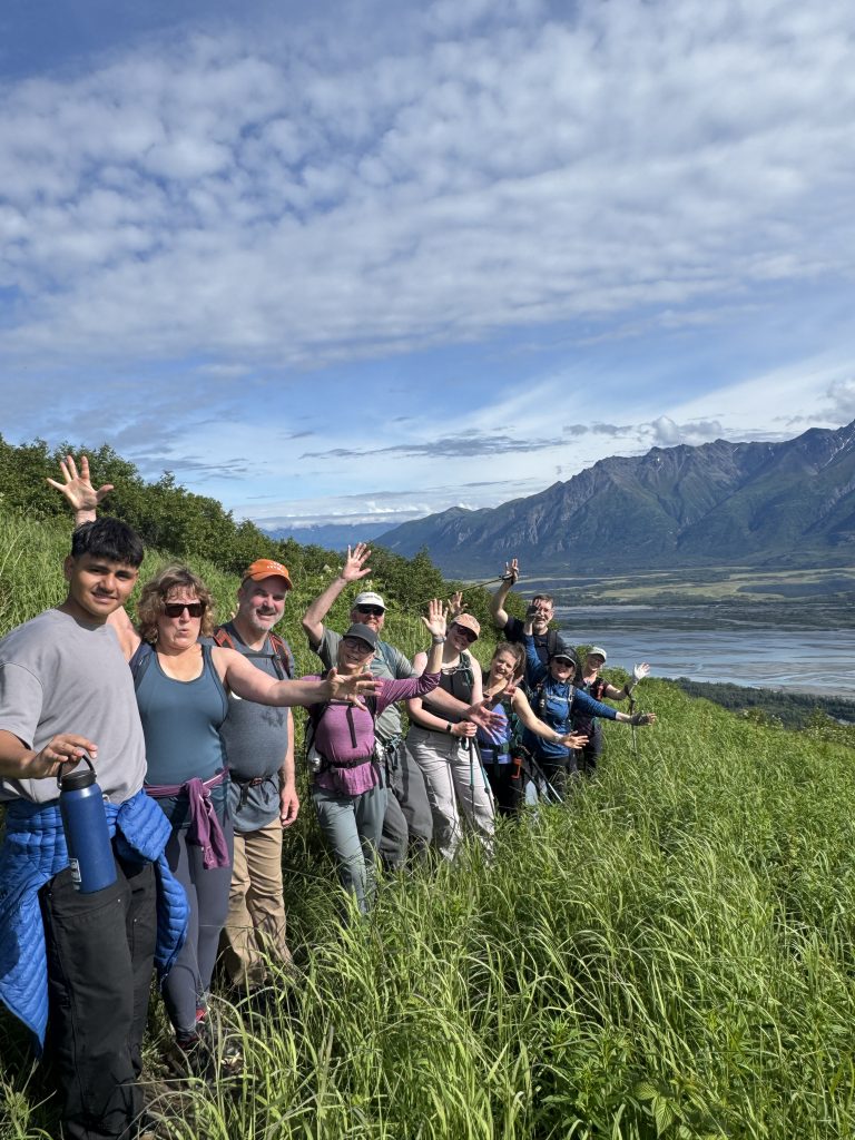 A group of hikers waving on a grassy hillside overlooking a valley and mountains in Mat-Su, Alaska