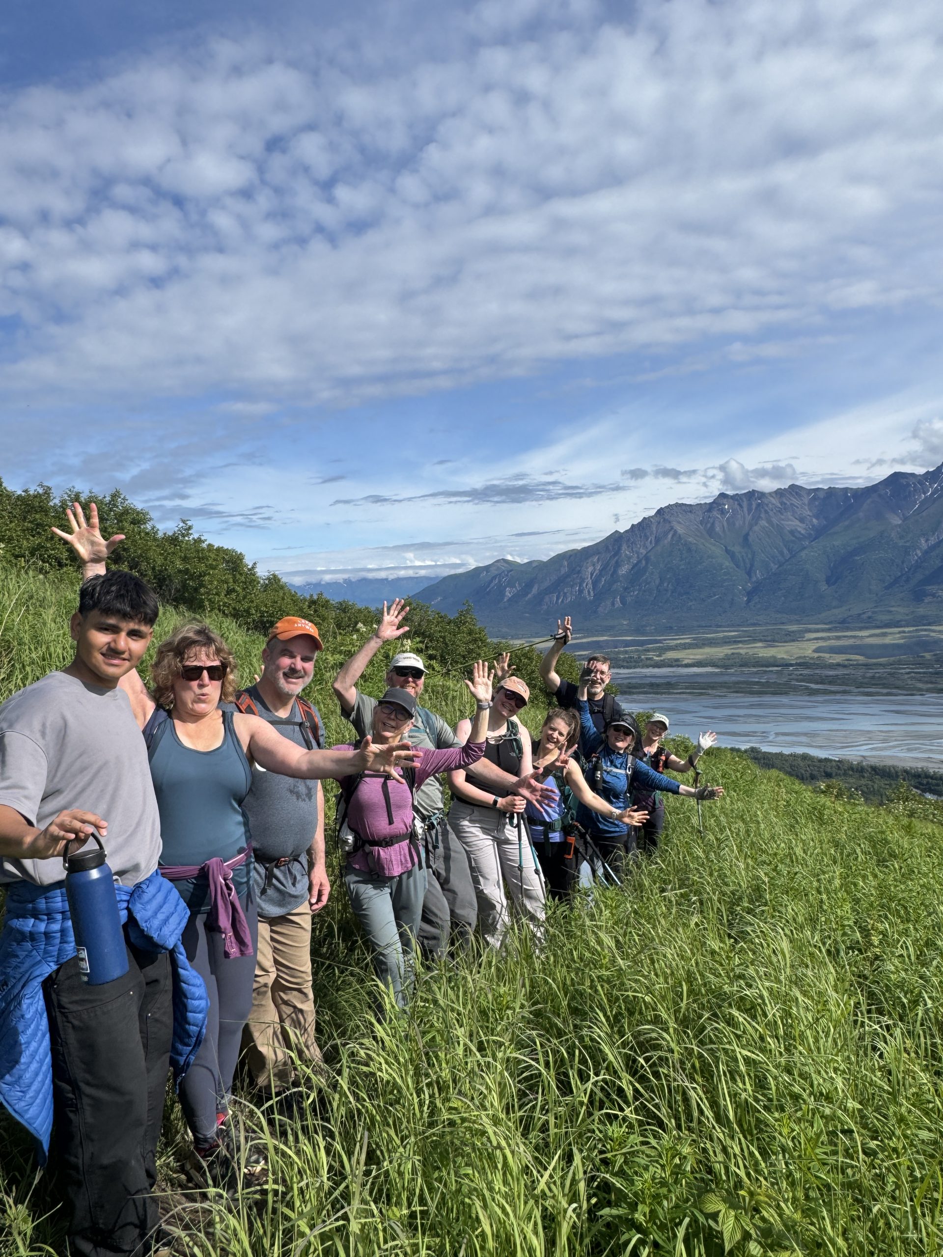 A group of hikers waving on a grassy hillside overlooking a valley and mountains in Mat-Su, Alaska