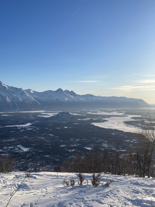A vast, snow-covered valley stretches beneath a range of jagged, snow-capped mountains in Mat-Su, Alaska