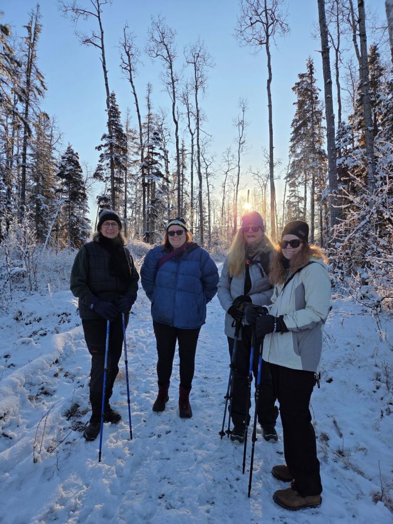 Four women standing on a snowy trail in a forest during sunset in Mat-Su, Alaska