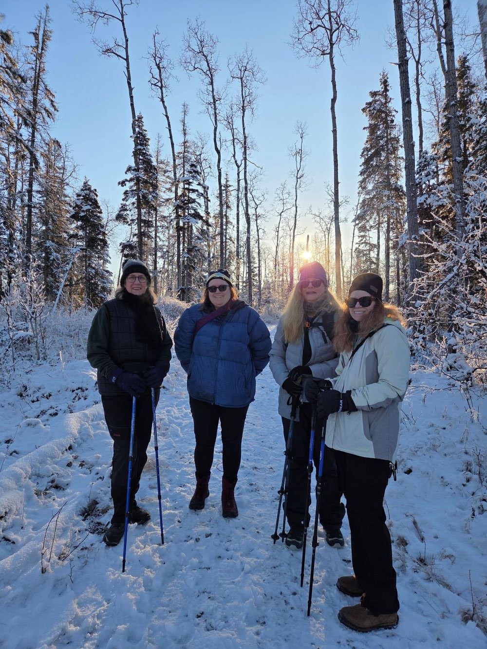 Four women standing on a snowy trail in a forest during sunset in Mat-Su, Alaska