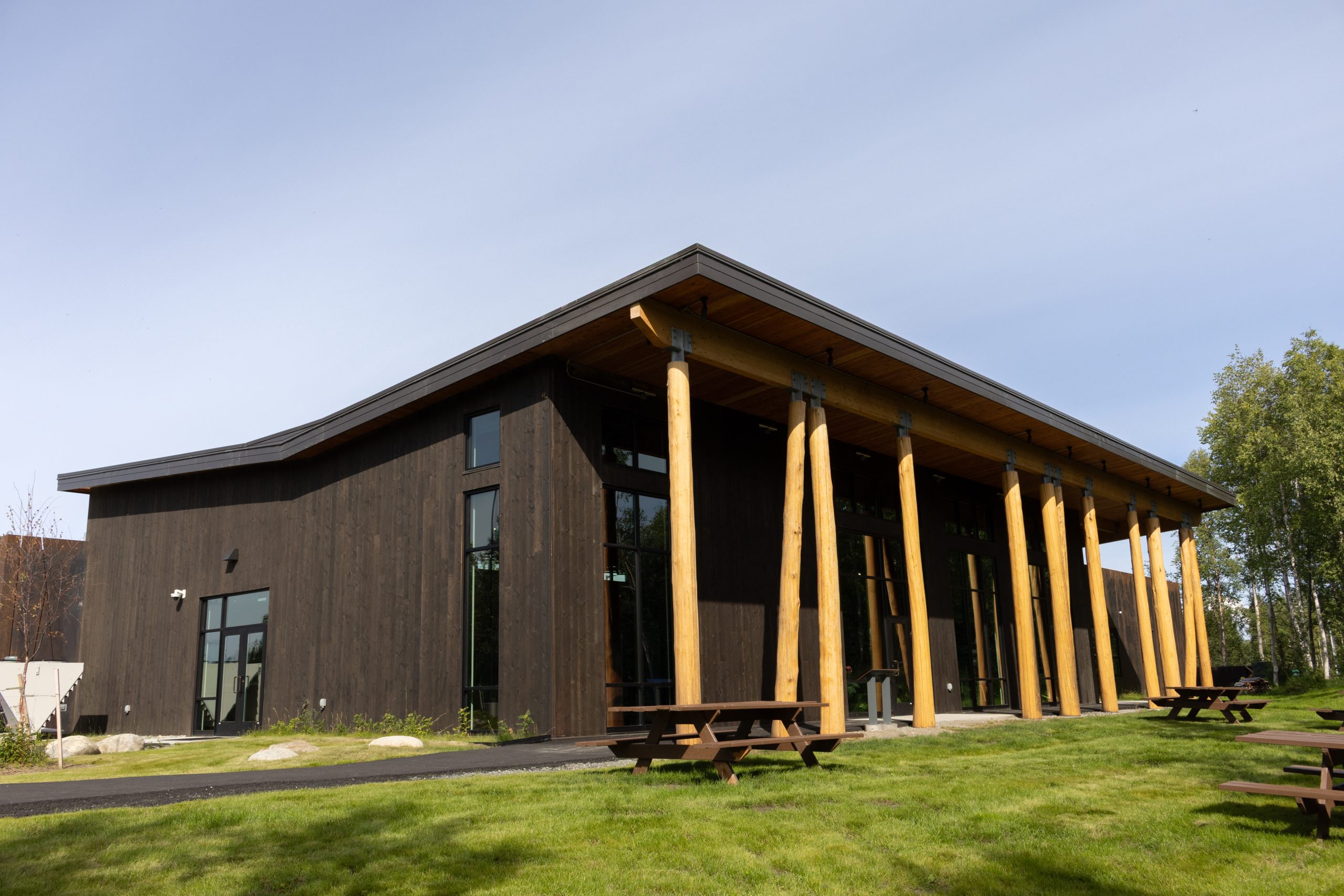 A modern dark wooden building with large log pillars and picnic tables on grass in Mat-Su, Alaska