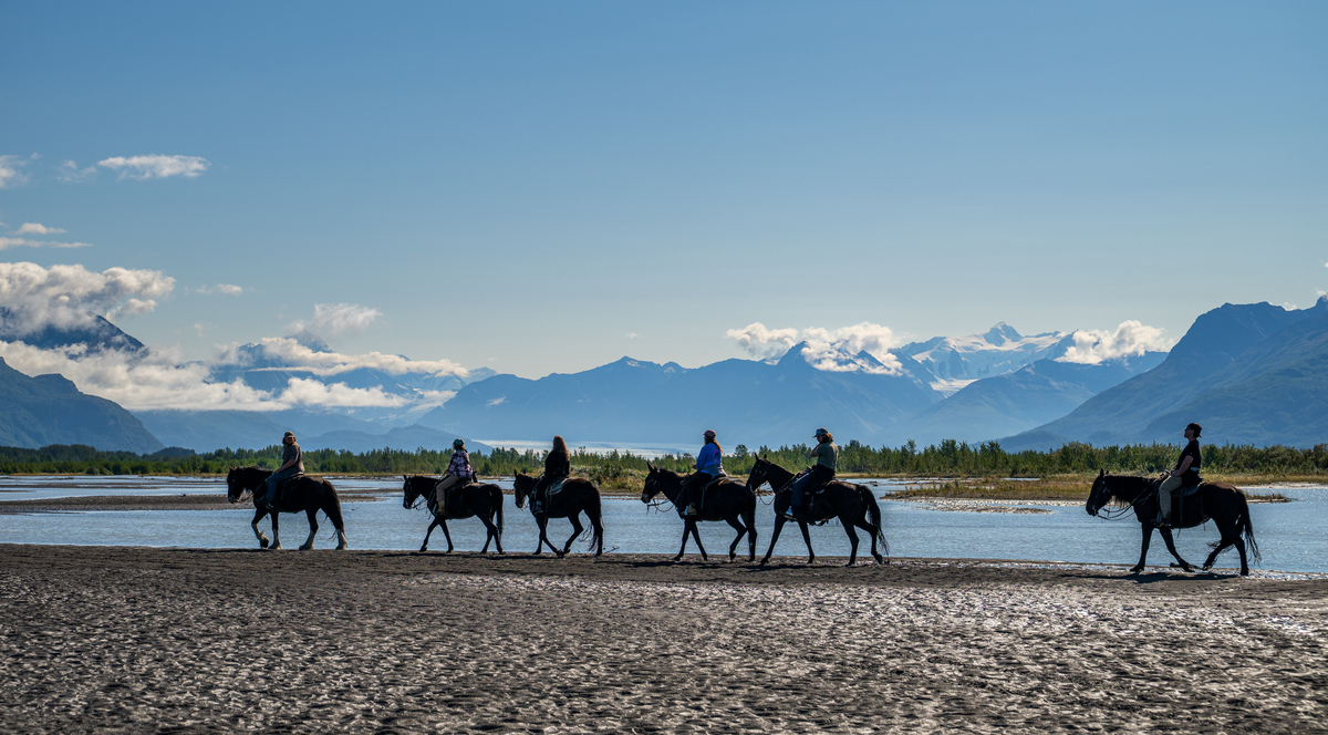 A group of people riding horses along a riverbank with mountains in the background in Mat-Su, Alaska